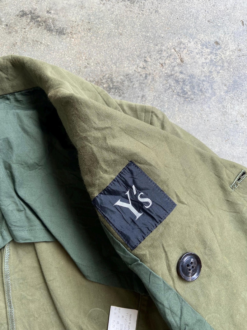 May include: Olive green jacket with a black label that reads "Y's" in white. The jacket has a button closure and a dark green lining. The fabric appears to be a durable, possibly cotton blend.