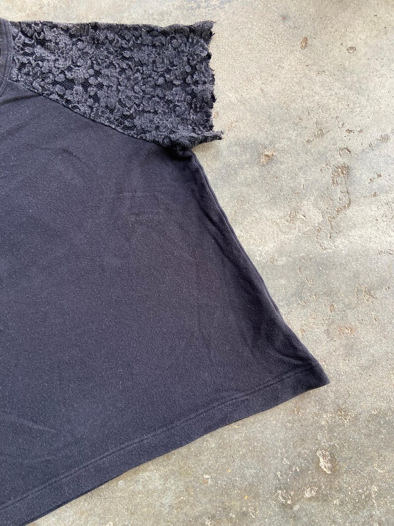 Pu&ograve; includere: T-shirt nero con dettaglio manica in pizzo strutturato. La manica corta &egrave; realizzata in pizzo scuro fantasia. Il resto della maglietta &egrave; di colore nero tinta unita.