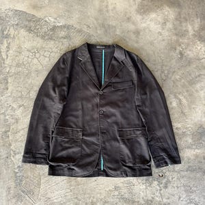 May include: A dark brown blazer featuring a notched lapel, two front buttons, and two large flap pockets. The garment includes a single chest pocket and a visible bright turquoise vertical trim detail along the inner front placket.