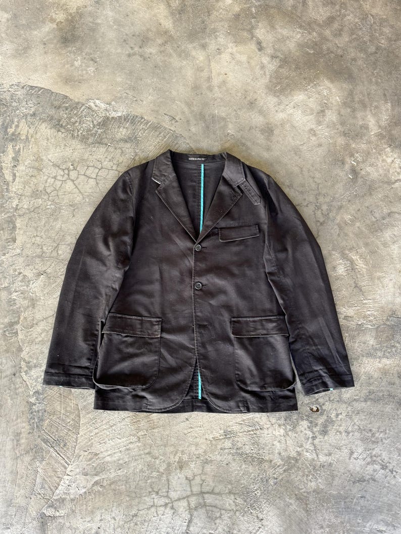 May include: A dark brown blazer featuring a notched lapel, two front buttons, and two large flap pockets. The garment includes a single chest pocket and a visible bright turquoise vertical trim detail along the inner front placket.
