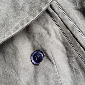 May include: Close-up of olive green fabric with a dark blue button. The button is sewn onto the fabric, and the fabric has a textured appearance. The image shows a portion of a garment, possibly a coat or jacket.