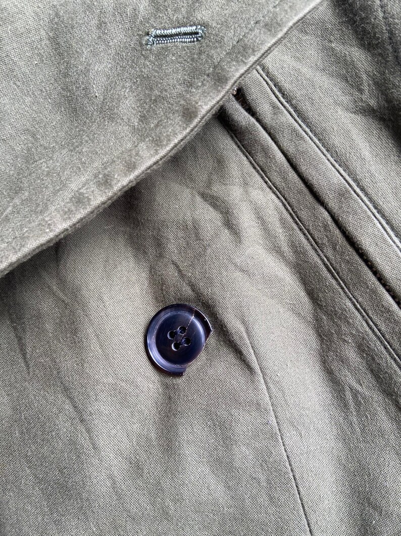 May include: Close-up of olive green fabric with a dark blue button. The button is sewn onto the fabric, and the fabric has a textured appearance. The image shows a portion of a garment, possibly a coat or jacket.