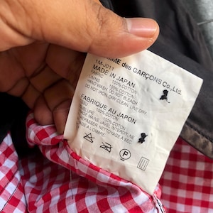 May include: A tag from a garment with text that reads "MADE IN JAPAN" and "100% COTTON". The tag is held up to show the red and white gingham lining of the garment. The brand name "...ME des GAR&Ccedil;ONS Co., Ltd." is visible.
