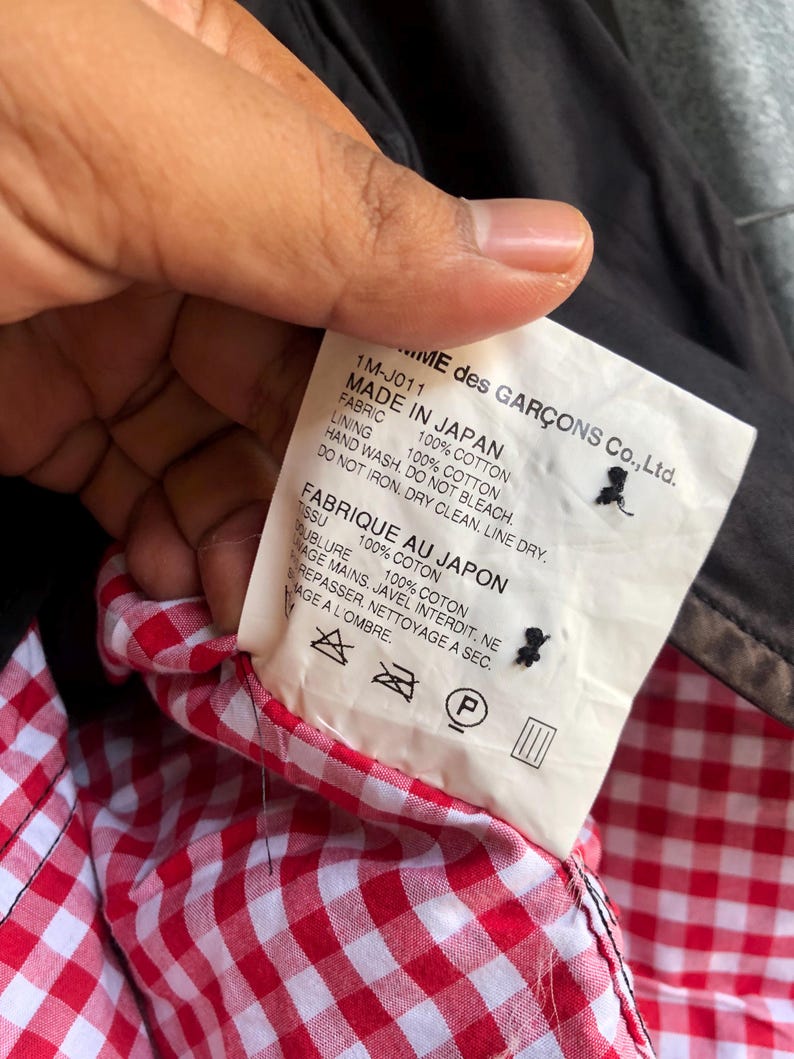 May include: A tag from a garment with text that reads "MADE IN JAPAN" and "100% COTTON". The tag is held up to show the red and white gingham lining of the garment. The brand name "...ME des GAR&Ccedil;ONS Co., Ltd." is visible.