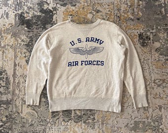 May include: A light beige crew neck sweatshirt featuring the text "U.S. ARMY AIR FORCES" in navy blue. The design includes a stylized emblem of wings. The sweatshirt has long sleeves and ribbed cuffs and hem.