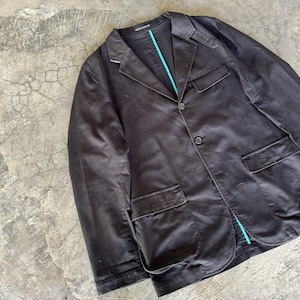 May include: A dark brown blazer featuring a notched lapel, two front buttons, and two large patch pockets. The garment includes a distinct bright turquoise interior lining visible along the front placket and collar area, with structured shoulder detailing.