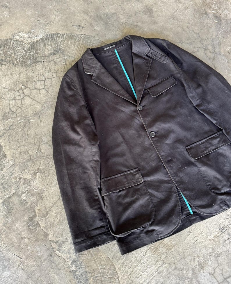 May include: A dark brown blazer featuring a notched lapel, two front buttons, and two large patch pockets. The garment includes a distinct bright turquoise interior lining visible along the front placket and collar area, with structured shoulder detailing.