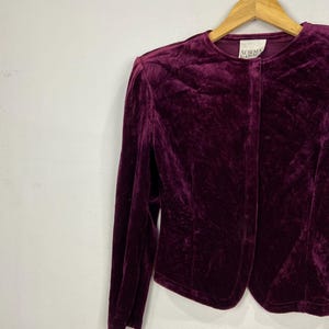 以下が含まれることがあります： 深紫色のベルベットのブレザーで、ボタンが1つ付いています。ブレザーは木製ハンガーにかけられています。襟には「Norma Kamali」のラベルが付いています。