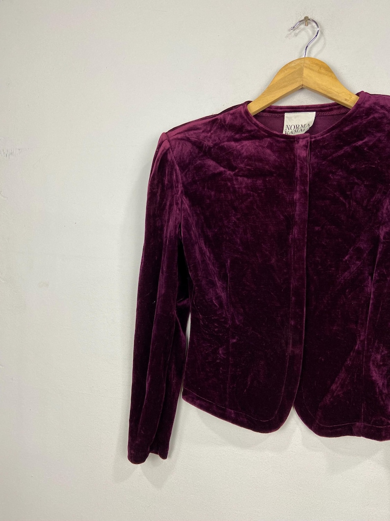 以下が含まれることがあります： 深紫色のベルベットのブレザーで、ボタンが1つ付いています。ブレザーは木製ハンガーにかけられています。襟には「Norma Kamali」のラベルが付いています。