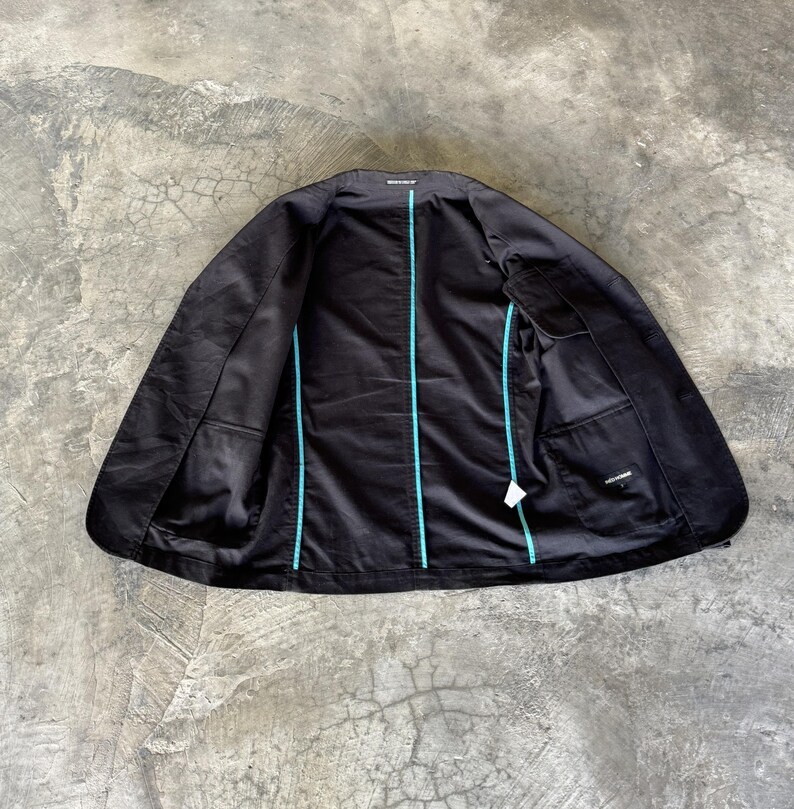 May include: A black blazer shown from the inside, featuring a solid black lining with bright turquoise piping along the seams. The garment includes an interior chest pocket on the right side and a small white care label attached to the lower seam.