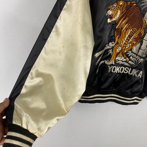 Puede incluir: Chaqueta de sat&eacute;n negra con manga color crema y ribete negro. La chaqueta presenta un dise&ntilde;o de tigre bordado con la palabra "Japan" y "Yokosuka" en escritura blanca. El pu&ntilde;o es a rayas negras y crema.