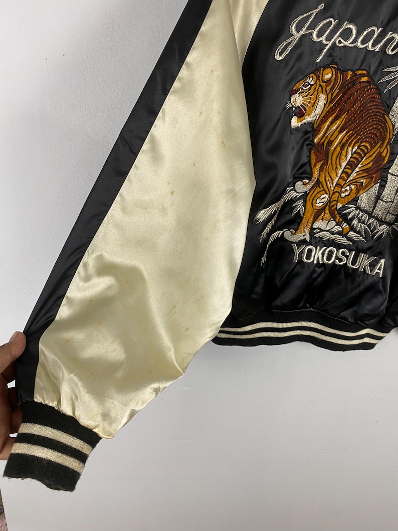 Puede incluir: Chaqueta de sat&eacute;n negra con manga color crema y ribete negro. La chaqueta presenta un dise&ntilde;o de tigre bordado con la palabra "Japan" y "Yokosuka" en escritura blanca. El pu&ntilde;o es a rayas negras y crema.