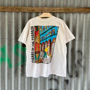 May include: White t-shirt with a colorful graphic on the back featuring an Egyptian-themed illustration. The graphic includes figures, hieroglyphs, and the text "CIGARS OF THE PHARAOH" vertically on the left side. The shirt is hanging on a hanger.