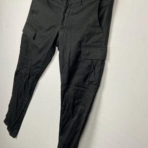 May include: Black cargo trousers with two side pockets and a button fastening. The trousers are hanging on a wooden hanger against a white wall. The trousers have a subtle herringbone pattern.
