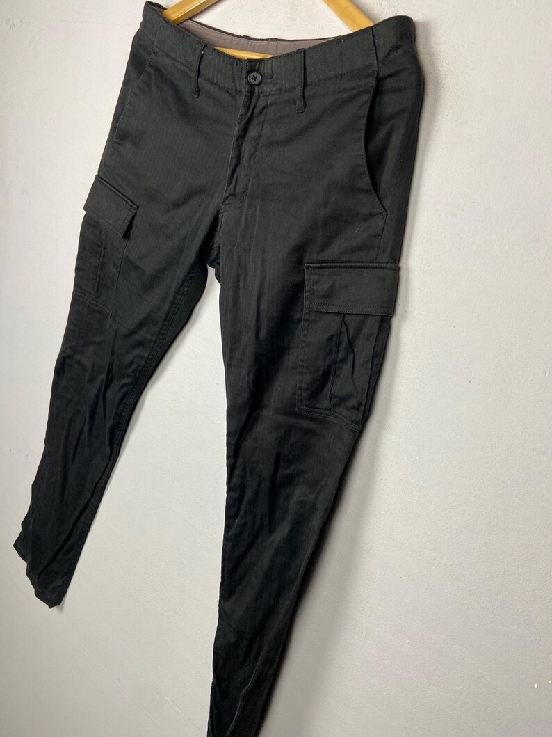 May include: Black cargo trousers with two side pockets and a button fastening. The trousers are hanging on a wooden hanger against a white wall. The trousers have a subtle herringbone pattern.