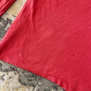 May include: A close-up of a red long-sleeved shirt. The shirt is made of a soft material and has a floral design on the upper right side. The shirt is slightly wrinkled.