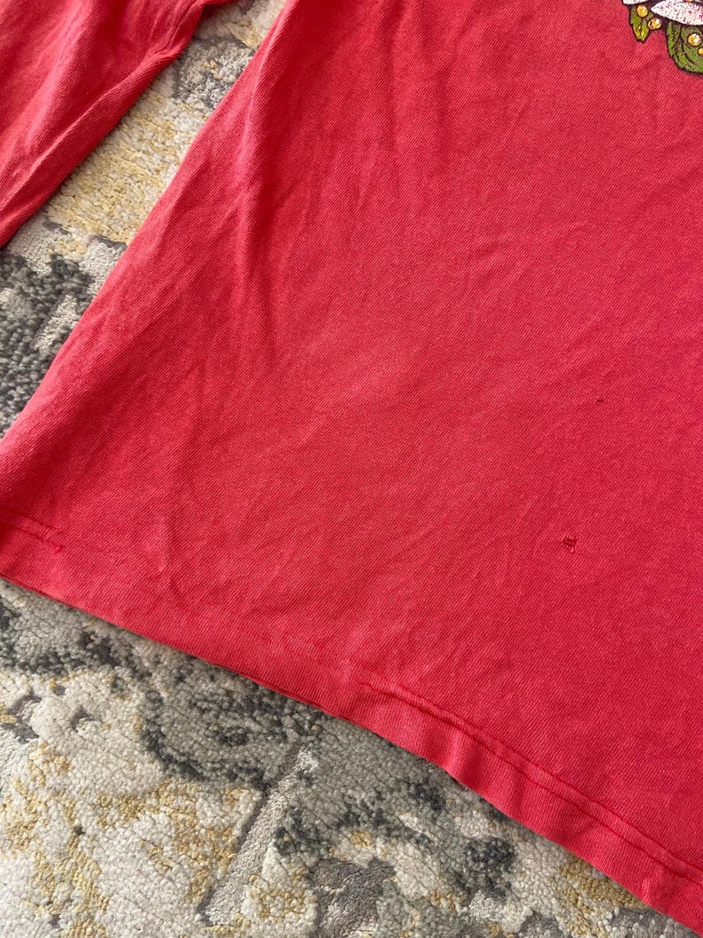 May include: A close-up of a red long-sleeved shirt. The shirt is made of a soft material and has a floral design on the upper right side. The shirt is slightly wrinkled.