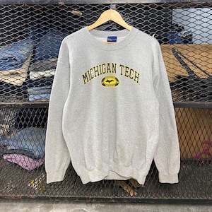 Puede incluir: Sudadera gris con un logotipo de Michigan Tech amarillo y negro. El logotipo dice "Michigan Tech" en un arco sobre un murciélago negro con alas amarillas y las palabras "Huskies Hockey" debajo.