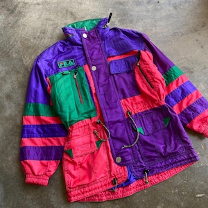 May include: A retro Fila jacket in a striking colour block design. The jacket features a mix of purple, green, and pink panels with striped accents. It has multiple pockets, a zip-up front, and the Fila logo patch.