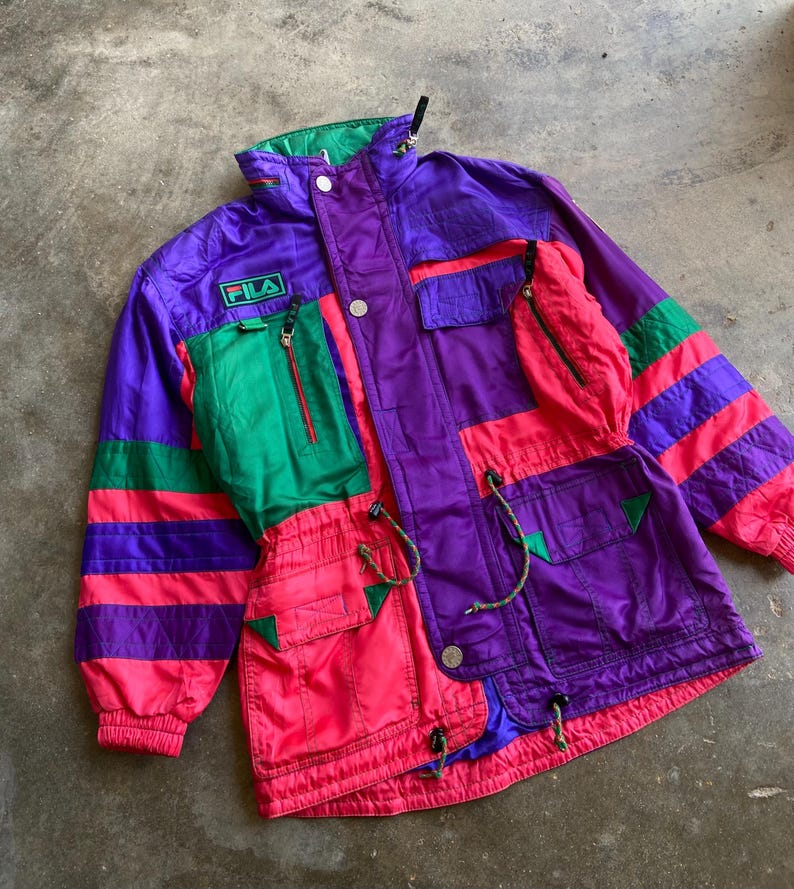 May include: A retro Fila jacket in a striking colour block design. The jacket features a mix of purple, green, and pink panels with striped accents. It has multiple pockets, a zip-up front, and the Fila logo patch.
