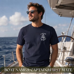 May include: A navy blue short-sleeved t-shirt featuring a white graphic on the left chest. The graphic displays a nautical anchor design with the text "CAPTAIN" arched above the anchor and the name "MARTIN" printed horizontally across the center.
