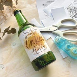 Könnte beinhalten: Eine grüne Bierflasche mit goldenem Verschluss und einem individuellen Etikett mit der Aufschrift "Gratulation Du Wirst Onkel Gotti". Eine weiße Schere und ein blauer Klebebandabroller liegen neben der Flasche.