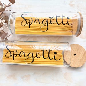 Könnte beinhalten: Zwei zylindrische Vorratsgläser aus klarem Glas, gefüllt mit langen gelben Spaghetti. Jedes Glas hat einen Bambusdeckel und eine schwarze kursive Aufschrift Spagötti und Spagotti mit kleinen Herzakzenten auf dem Glas und dem Deckel.