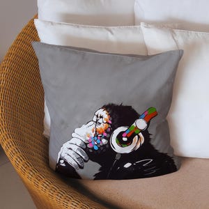 May include: Gray square throw pillow featuring a colorful illustration of a chimpanzee wearing headphones. The chimpanzee is depicted in a graffiti art style, with vibrant splashes of color on its face and the headphones. The pillow is placed on a wicker chair.
