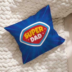 May include: A blue decorative pillow with a comic book style design. The pillow features a red, white, and yellow emblem that reads "SUPER DAD". The background is blue with radial lines and a dotted pattern.
