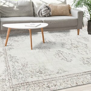 Puede incluir: Una alfombra blanca con un diseño vintage y desgastado. La alfombra tiene un patrón sutil de motivos florales descoloridos y un borde con un diseño más intrincado. La alfombra se muestra en un ambiente de sala de estar con dos sofás y una mesa de café.