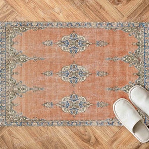 May include: A vintage-style rug with a faded orange background and intricate blue and green patterns. Two white slippers are placed on the rug.