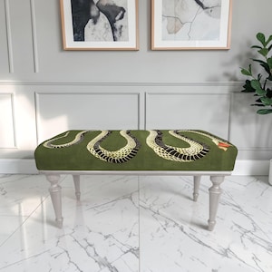May include: A rectangular bench featuring a dark olive green upholstered seat with a large, winding snake pattern in black and yellow. The snake has a small red accent near its head. The bench is supported by four light gray, tapered wooden legs.