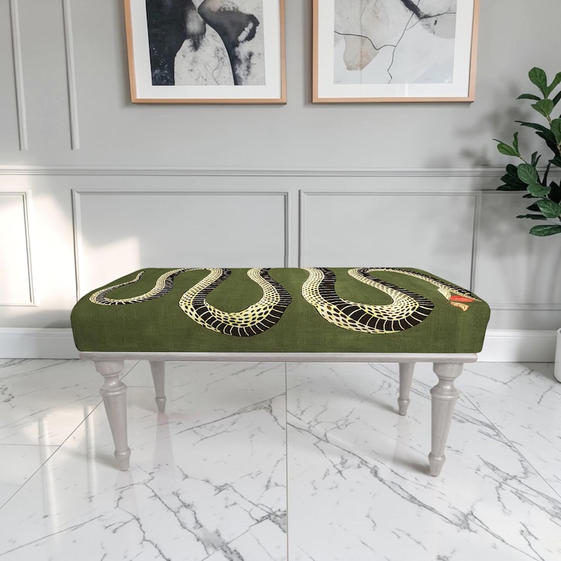 May include: A rectangular bench featuring a dark olive green upholstered seat with a large, winding snake pattern in black and yellow. The snake has a small red accent near its head. The bench is supported by four light gray, tapered wooden legs.