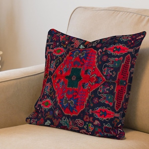 May include: A decorative pillow with a traditional rug design. The pillow features a central red medallion with intricate blue, green, and gold patterns against a dark navy background. The pillow is placed on a beige sofa.