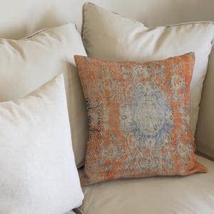 Peut inclure: Un coussin à motifs orange et bleu repose sur un canapé crème, avec deux autres coussins crème en arrière-plan. Le coussin à motifs a un aspect vintage et vieilli.