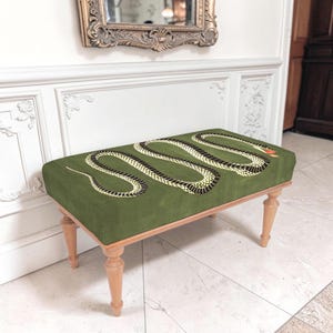 May include: A rectangular bench featuring a deep olive green velvet upholstered top with a large, winding black and yellow snake graphic printed across the surface. The bench is supported by four light-toned wooden legs with a turned spindle design.
