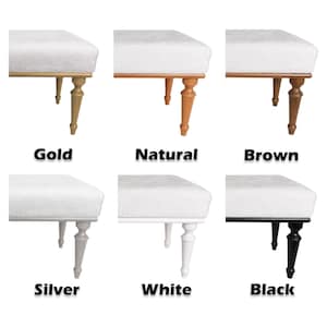 May include: A collection of six ottomans featuring white upholstered tops and turned legs in various finishes labeled as Gold, Natural, Brown, Silver, White, and Black. Each ottoman displays a consistent design with a rectangular seat and four legs.