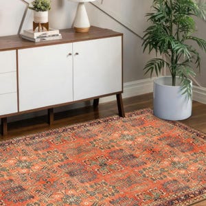 May include: An orange and blue patterned area rug with a dark border. The rug is placed on a wooden floor. A white cabinet with a wooden top and a white lamp are in the background.