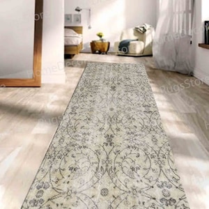 Puede incluir: Una alfombra rectangular larga con fondo beige y un patrón floral gris. La alfombra está colocada sobre un suelo de madera en una habitación con paredes blancas.