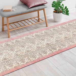 Puede incluir: Un tapete de corredor de color rosa y crema con un borde rosa. El tapete está colocado sobre un suelo de madera clara delante de un banco de madera con un cojín gris.