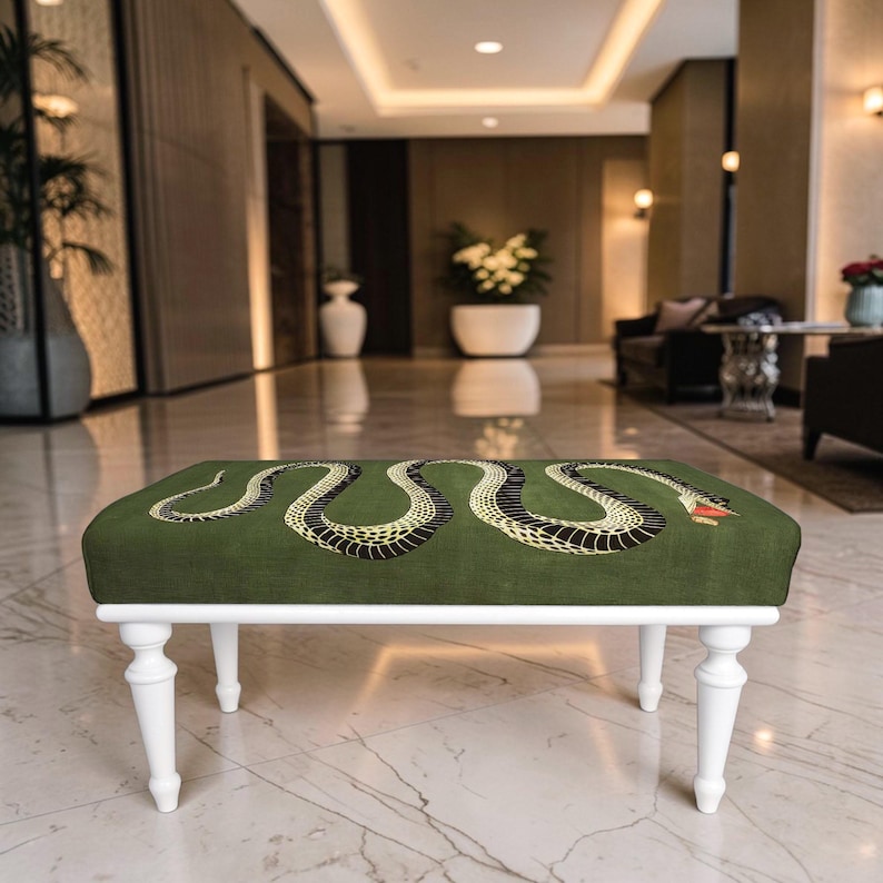May include: A rectangular ottoman featuring a dark olive green upholstered top with a large, detailed black and yellow snake graphic across the surface. The piece is supported by four white, turned wooden legs in a classic spindle design.