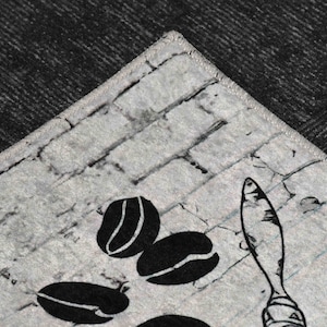 May include: A close-up of a gray and white kitchen rug with a brick pattern. The rug features a black and white illustration of coffee beans and a spoon.