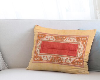 May include: A rectangular decorative pillow with a yellow background and a central red rectangle. The pillow features an ornate border with floral and geometric patterns in shades of orange and white. The pillow is resting on a white sofa.