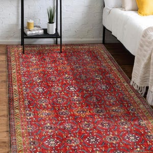 May include: A rectangular, red oriental rug with a detailed floral pattern. The rug has a border with a mix of colors including gold, green, and blue. The rug is placed on a wooden floor, near a bed and a black shelf.