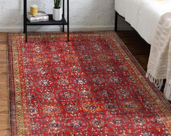 May include: A rectangular, red oriental rug with a detailed floral pattern. The rug has a border with a mix of colors including gold, green, and blue. The rug is placed on a wooden floor, near a bed and a black shelf.