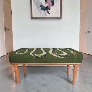 May include: A rectangular bench with four light brown wooden legs and a green upholstered seat. The seat features a large, winding black and yellow snake graphic with a red mouth, stretching across the entire length of the fabric surface.