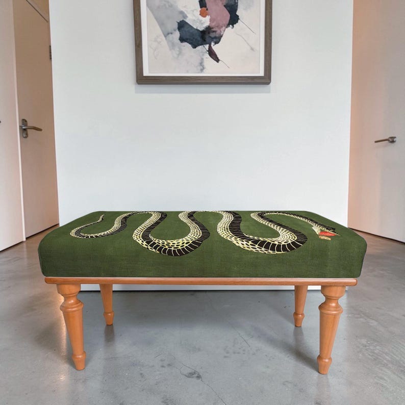 May include: A rectangular bench with four light brown wooden legs and a green upholstered seat. The seat features a large, winding black and yellow snake graphic with a red mouth, stretching across the entire length of the fabric surface.