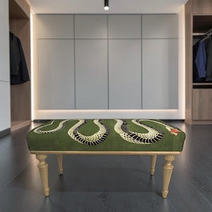 May include: A rectangular ottoman featuring a dark green upholstered top with a large, winding black and cream snake pattern across the surface. The piece is supported by four light-colored wooden legs with a turned, tapered design.