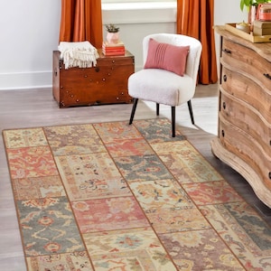 Può includere: Un tappeto rettangolare con un design patchwork. Presenta una varietà di colori, tra cui rosso, blu, beige e marrone. Il tappeto è posizionato su un pavimento in legno grigio chiaro. Sullo sfondo, una sedia bianca con gambe nere e un cuscino rosa.