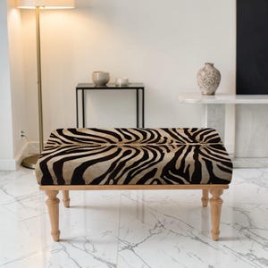 Könnte beinhalten: Ein rechteckiger Hocker mit einem Zebra-Print-Oberteil und hellen Holzbeinen. Der Zebra-Print hat schwarze und beige Streifen. Die Beine sind gedreht und haben ein klassisches Design. Der Hocker ist ein dekoratives Möbelstück.